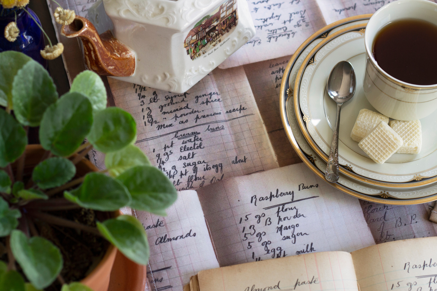 A table runner with a design of overlapping handwritten recipes in black cursive font lays down the center of a table. An off-white saucer with a tan rim and small navy accents sits to the right of the photo with a silver spoon to the left, three small square wafer cookies to the bottom and a cup of black tea in the center. A brown planter with green leaves on brown stems is to the left of the saucer. A cream-colored teapot with a brown spout sits at the top of the photo with a drawing of an old hotel.