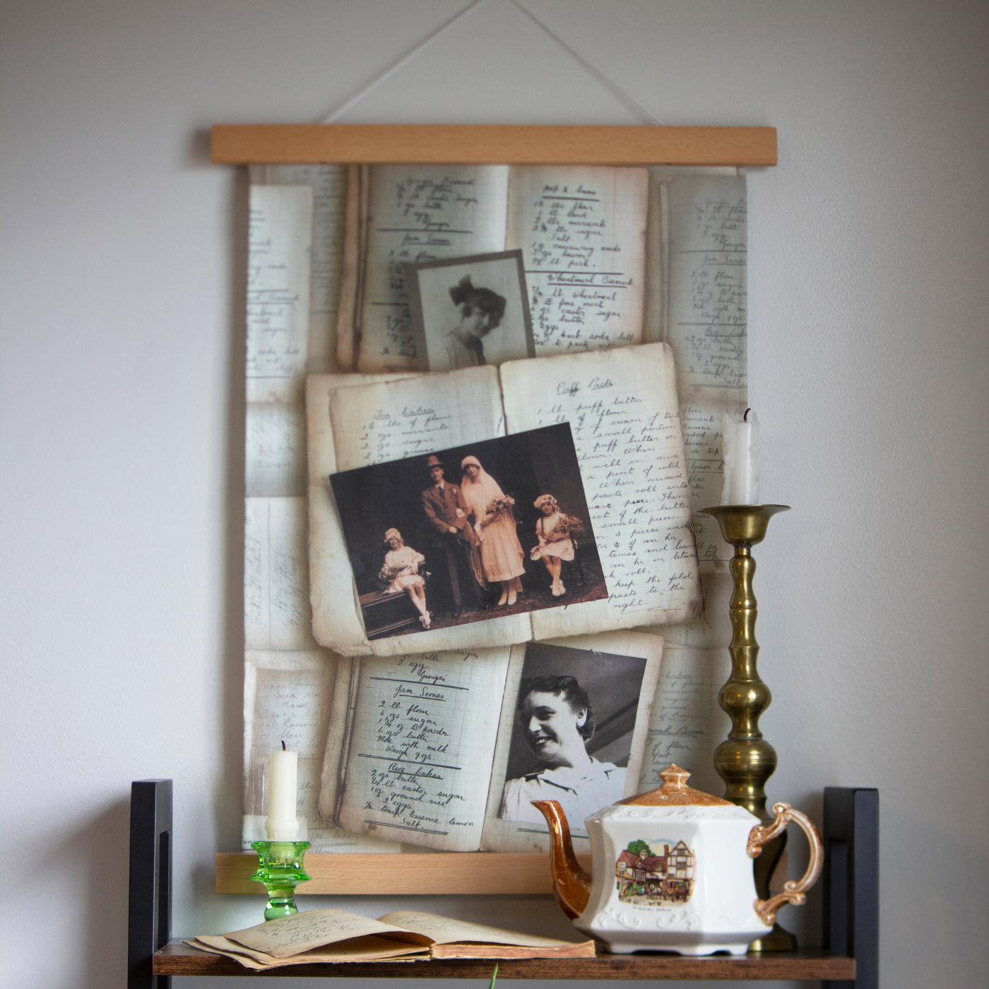 A wall hanging featuring handwritten recipes scattered along the back of the design and three photos on the top of the recipes, the top photo is a vintage photo of a woman smiling at the camera, the middle photo is a 1924 wedding photo with a groom and a bride standing in the middle of the photo with two small girls sitting at either side of them holding flower bouquets and the bottom photo is another vintage photo of a woman smiling and looking to the left. The wall hanging is hanging against a white wall and above a bookcase with brown shelves and black edges. A short green candlestick with a short white candle in it, an open book, a cream teapot with a brown top, handle and spout and a drawing on a hotel on it and a short white candle in a brass candlestick, are visible on the top shelf from left to right.