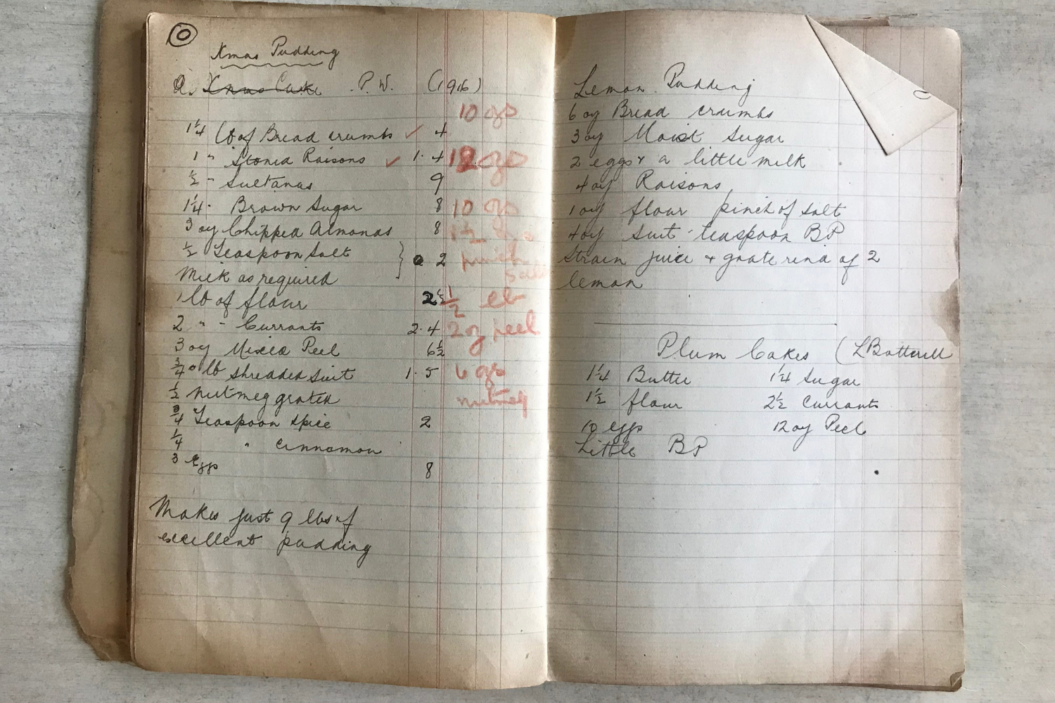 A handwritten recipe book lays open flat on a white table, showing both left and right pages of the book. The cursive text is written in thin black ink. On the lefthand page is a recipe for Xmas pudding. On the righthand page, is a recipe for lemon pudding at the top and plum cakes at the bottom.