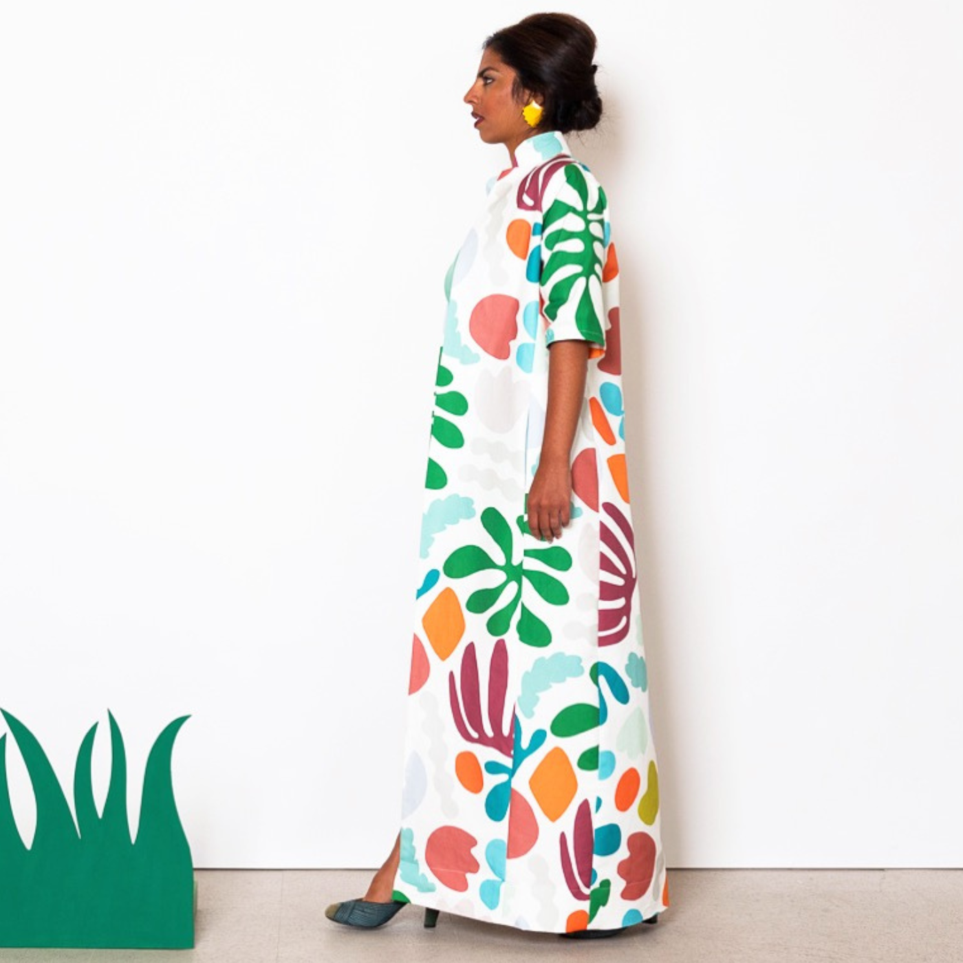A model stands facing to the left looking straight ahead off camera and wears a design with a white background featuring bright geometric shapes in green, turquoise, orange and dark red. A white wall is behind the model. A calf-high fake grass prop is to the photo’s left edge.