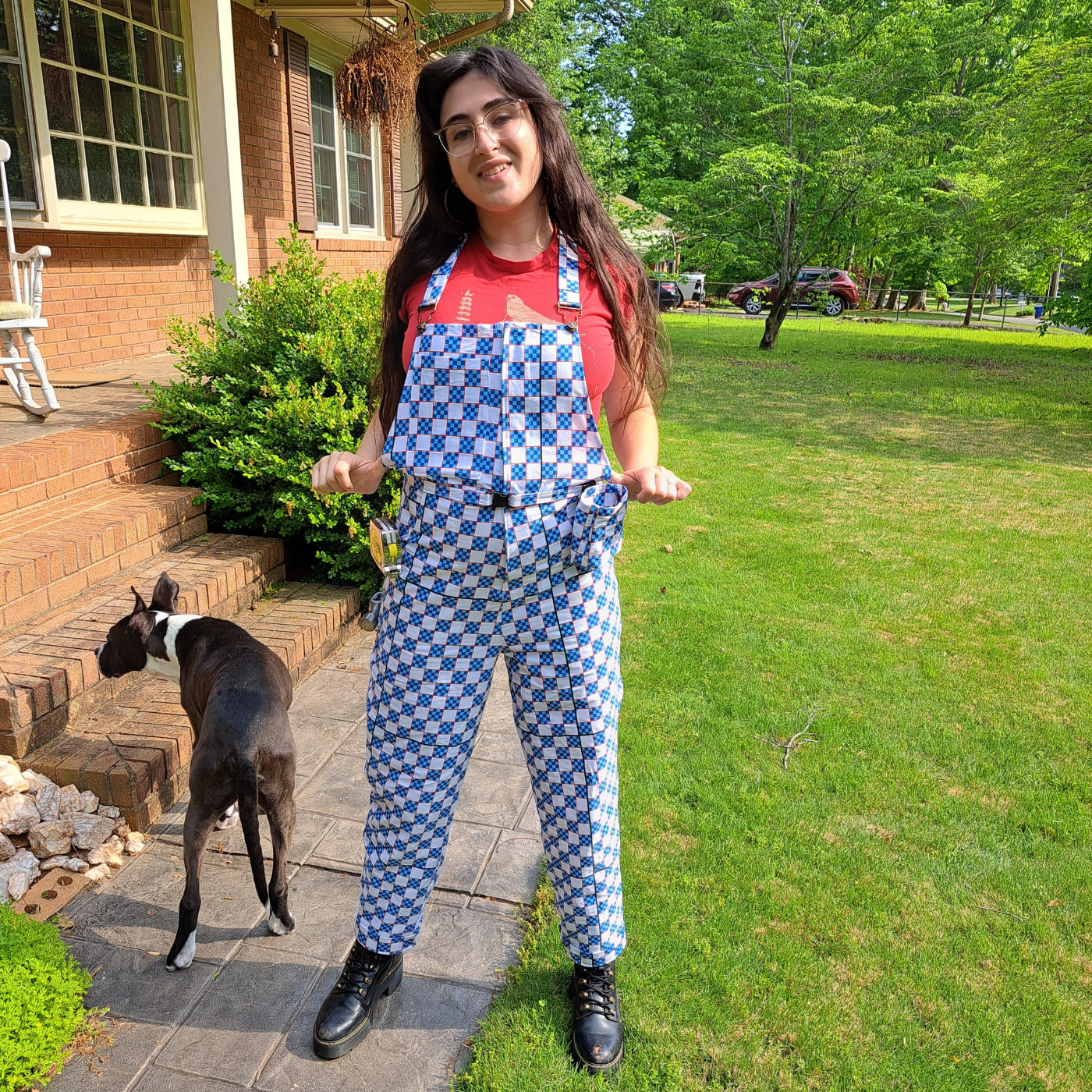Ace stands smiling in front of a red brick house in a grass yard wearing a pair of overalls in a print featuring a checkerboard design that also doubles as a ruler. The checkerboard is white and small-blue-and-navy blocks with all blocks outlined in red. A black dog with white accents is standing by their right foot.