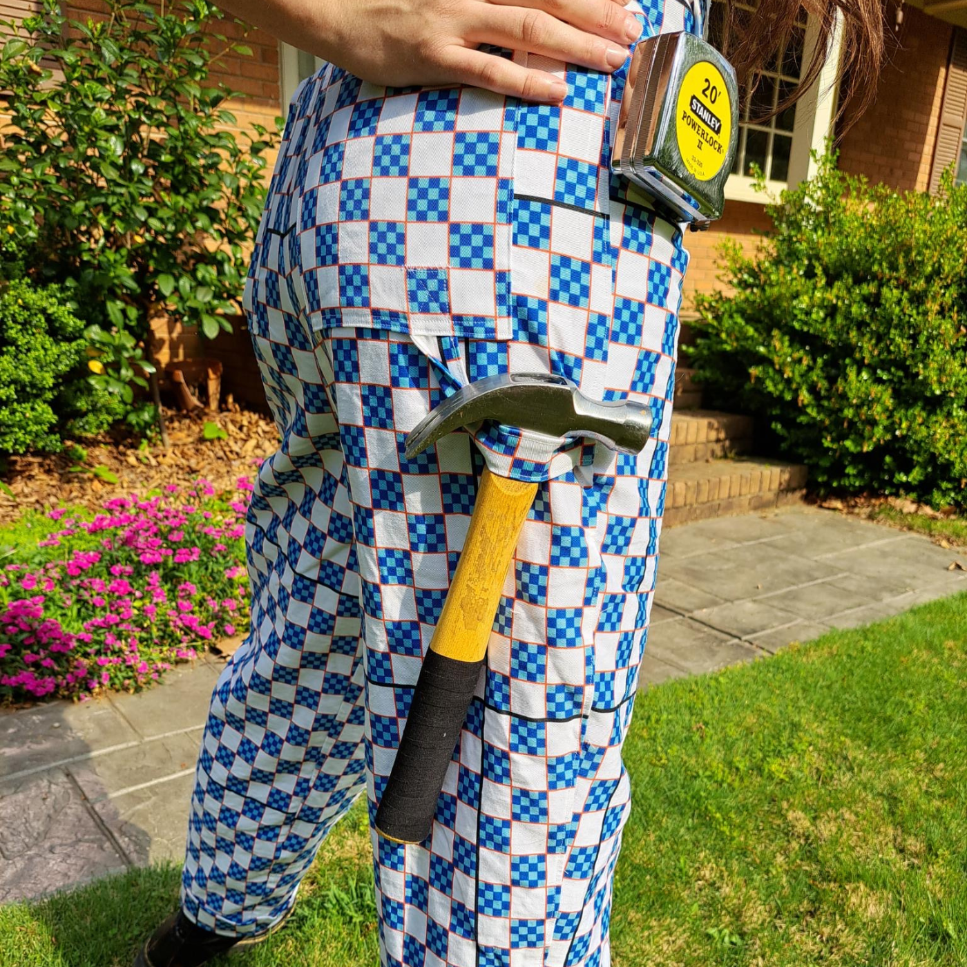 A close up of the back a pair of overalls in a print featuring a checkerboard design that also doubles as a ruler. The checkerboard is white and small-blue-and-navy blocks with all blocks outlined in red. A hammer hangs from a hammer loop under their right. Measuring tape in a metal case hangs up higher on the right side from a tool belt added to the garment.