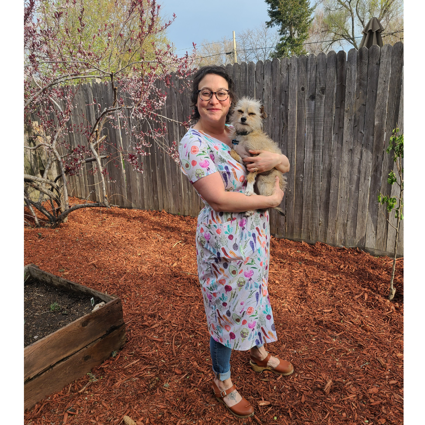 Angela stands looking at the camera and smiling and holding a small furry tan dog. She is wearing a dress with a cream background and small rainbow vegetables all over it. She is wearing the dress over pants and standing in the mulch as if in a backyard near a brown wooden fence.