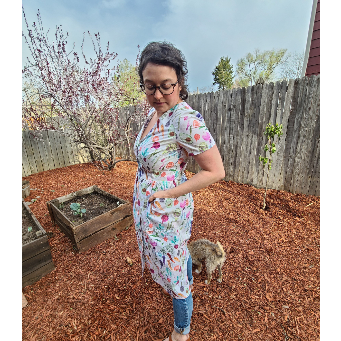 Angela stands to the side, looking down. A small furry tan dog is standing behind her. She is wearing a dress with a cream background and small rainbow vegetables all over it. She is wearing the dress over pants and standing in the mulch as if in a backyard near a brown wooden fence.
