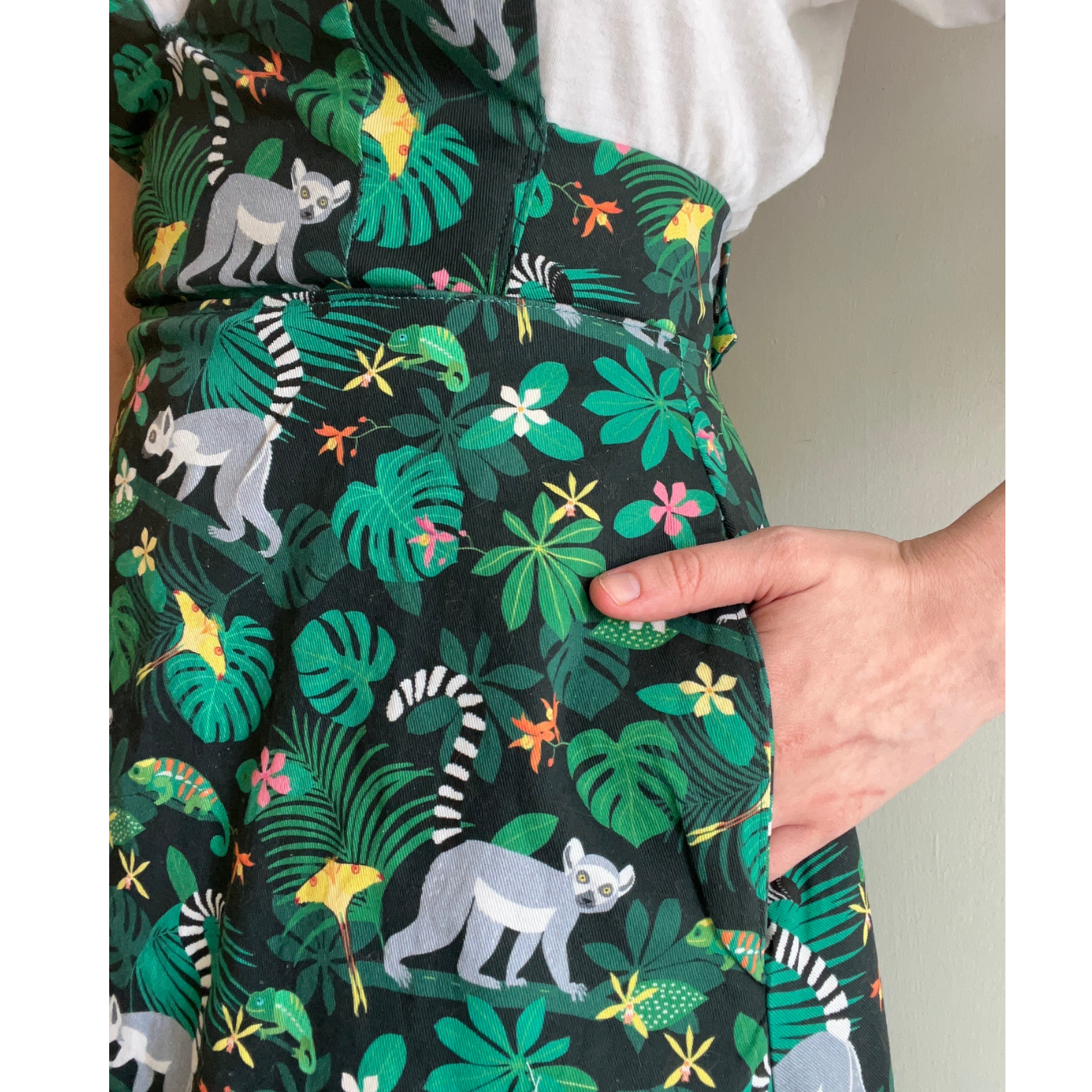 A close up of a pocket of the pinafore Anna is wearing with a black background and a repeating design with lemurs roaming around large green leaves with small white and yellow flowers. Her left hand is in her pocket.