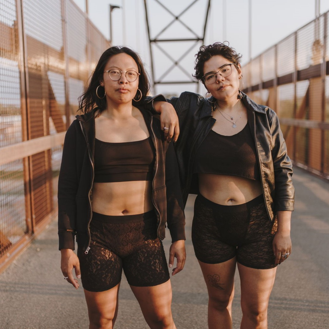 Two models are wearing cropped black tank tops and black lace boxer briefs. The model on the left is wearing a black hoodie and the model on the right is wearing a cropped black leather jacket. They are standing on a concrete bridge with metal sides.
