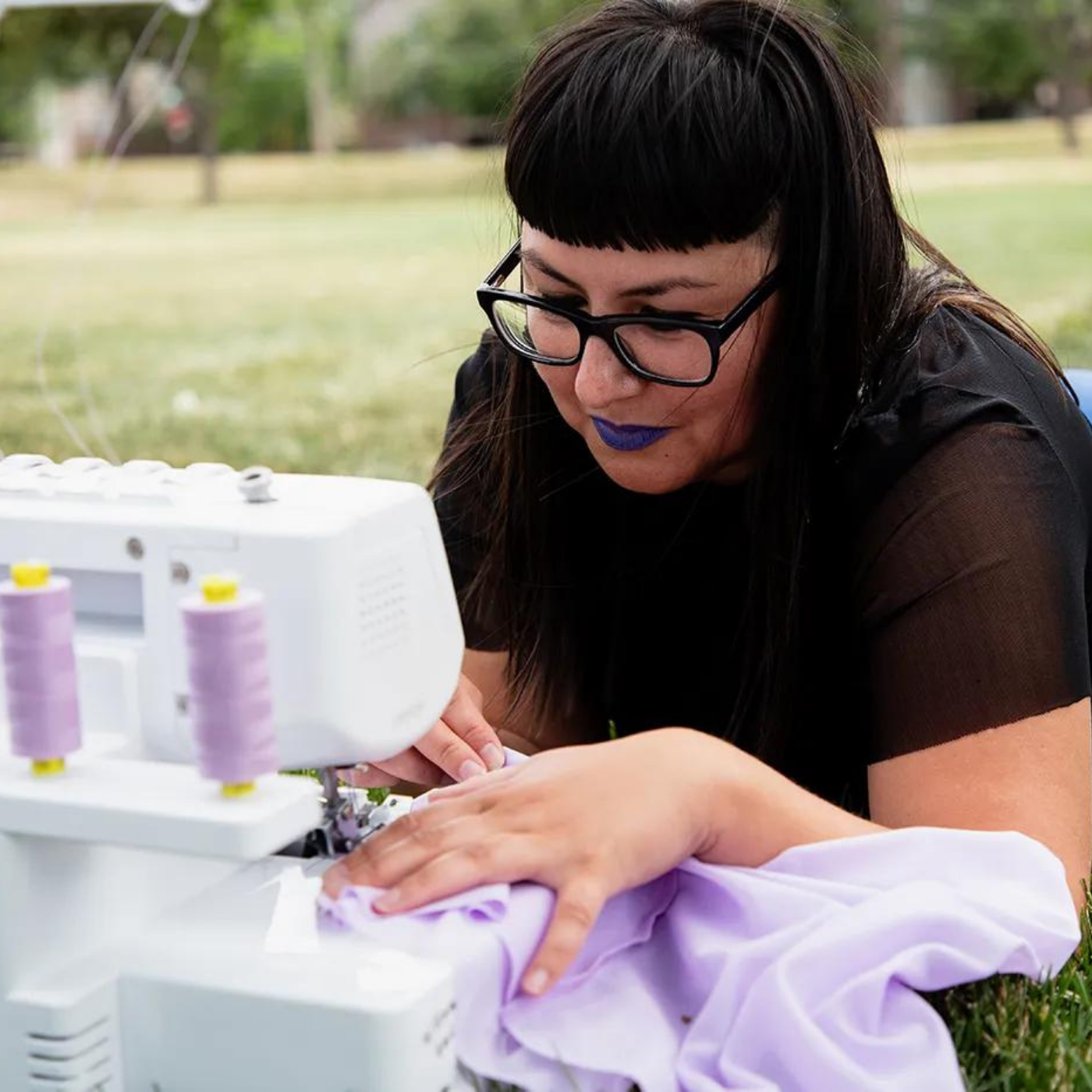 Mel Martinez sews purple fabric with two spools of purple thread visible on a serger. Mel is laying down in the grass with the sewing machine in front of her.