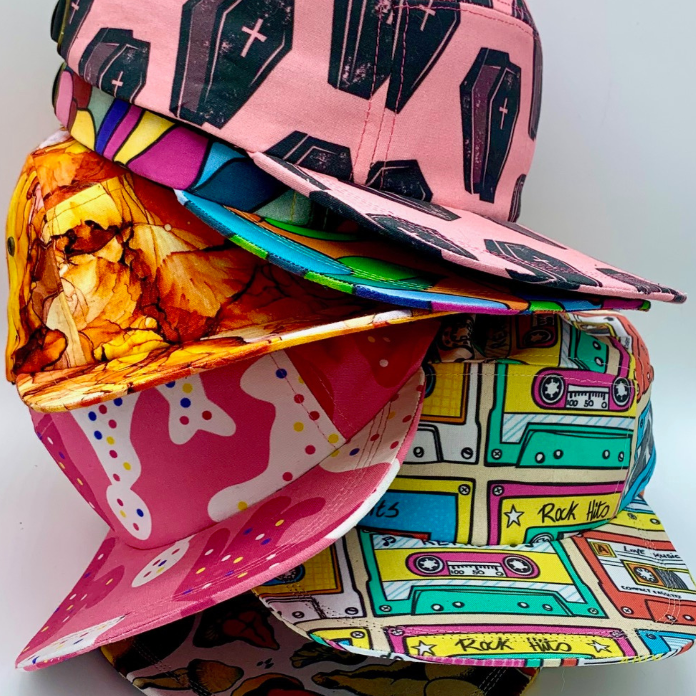 A stack of six baseball caps. From top to bottom, their featured designs are black coffins on a pink background; multicolored geometric jewel tone blocks; orange, black and yellow swirls; white blobs with red, yellow and blue small dots on a hot pink background; cassette tapes, some green, some orange, some yellow on a cream background; and in the shadow of the previous hat, what appears to be mushroom designs and a white background.