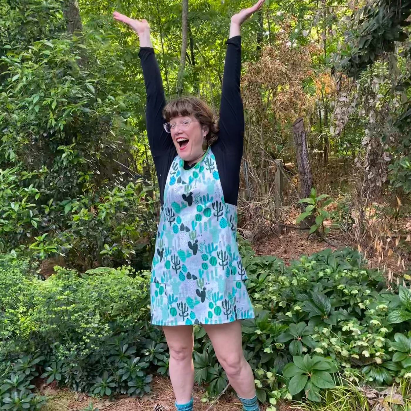 Betsy smiles and is wearing a pinafore with a gray background and repeating design of differing types of cacti. She is standing in a backyard full of green plants. Her hands are held up straight over her head.