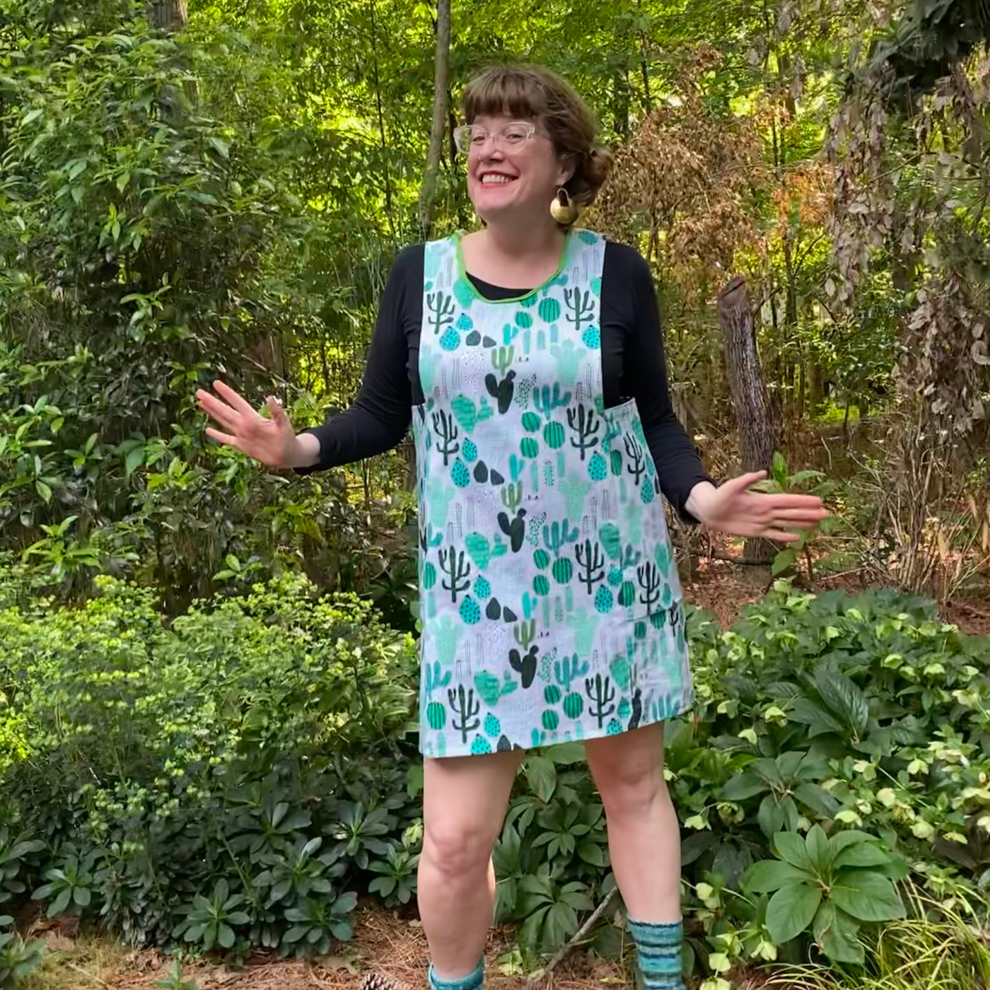 Betsy smiles and is wearing a pinafore with a gray background and repeating design of differing types of cacti. She is standing in a backyard full of green plants. Her hands are bent at the elbow and held out by her waist.