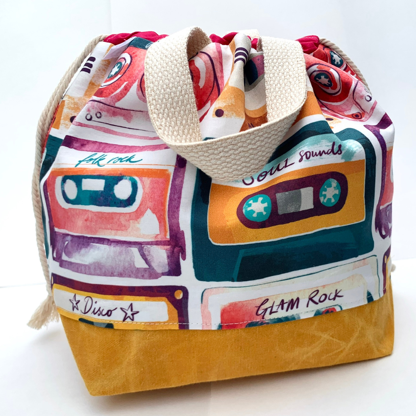 A fabric drawstring bag featuring a print with rows of watercolor cassette tapes, each titled a different 1970s rock genre, including disco, glam rock, soul sounds, folk rock, etc., and in a different muted jewel tone color, some yellow, some dark peach, some red, some dark green. The bag has a yellow fabric waxed bottom along with cream fabric handles and a drawstring closure.