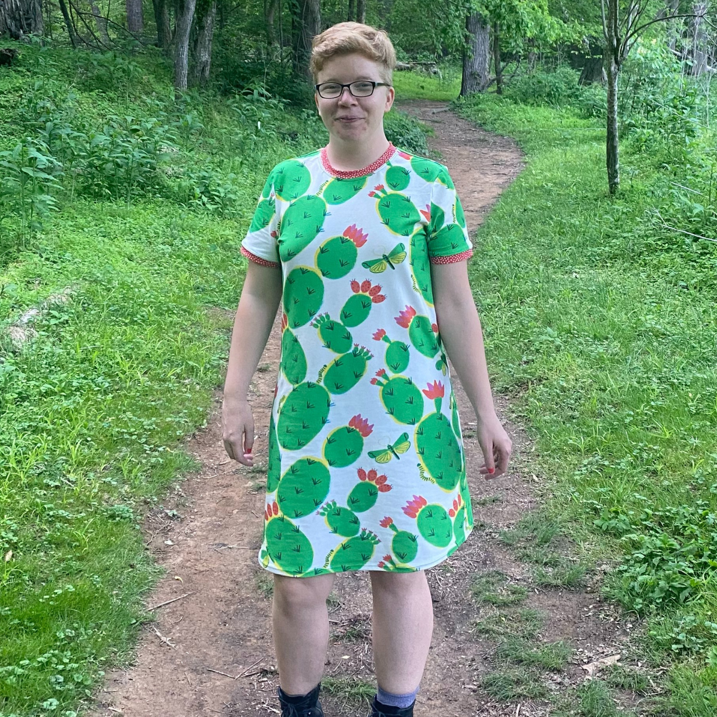 Bri stands in the middle of a dirt path going through the woods behind her. She is smiling at the camera and wearing a short-sleeved dress with a white background and a repeating green cactus design. The cacti have red blossoms and green-and-yellow moths floats near some of the blossoms. The sleeves and neckline have been ringed with red bias tape that has white dots.
