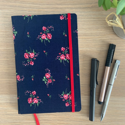 A notebook with a floral exterior (featuring a navy background, columns of small pink flowers with small green leaves and a red elastic closure and bookmark sits on a wooden desk next to three pens.