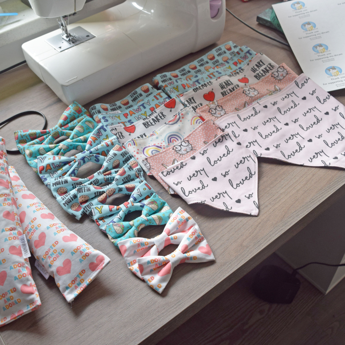 Cat kickers, bowties and bandanas lay in neat rows on a wooden table in front of a sewing machine. The accessories are in pink, blue and white fabrics, including a design with pink hearts that says “Adopted and adored” in rainbow pastel letters and a design with red hearts and the words “Heart Breaker” written in black all caps, among them.