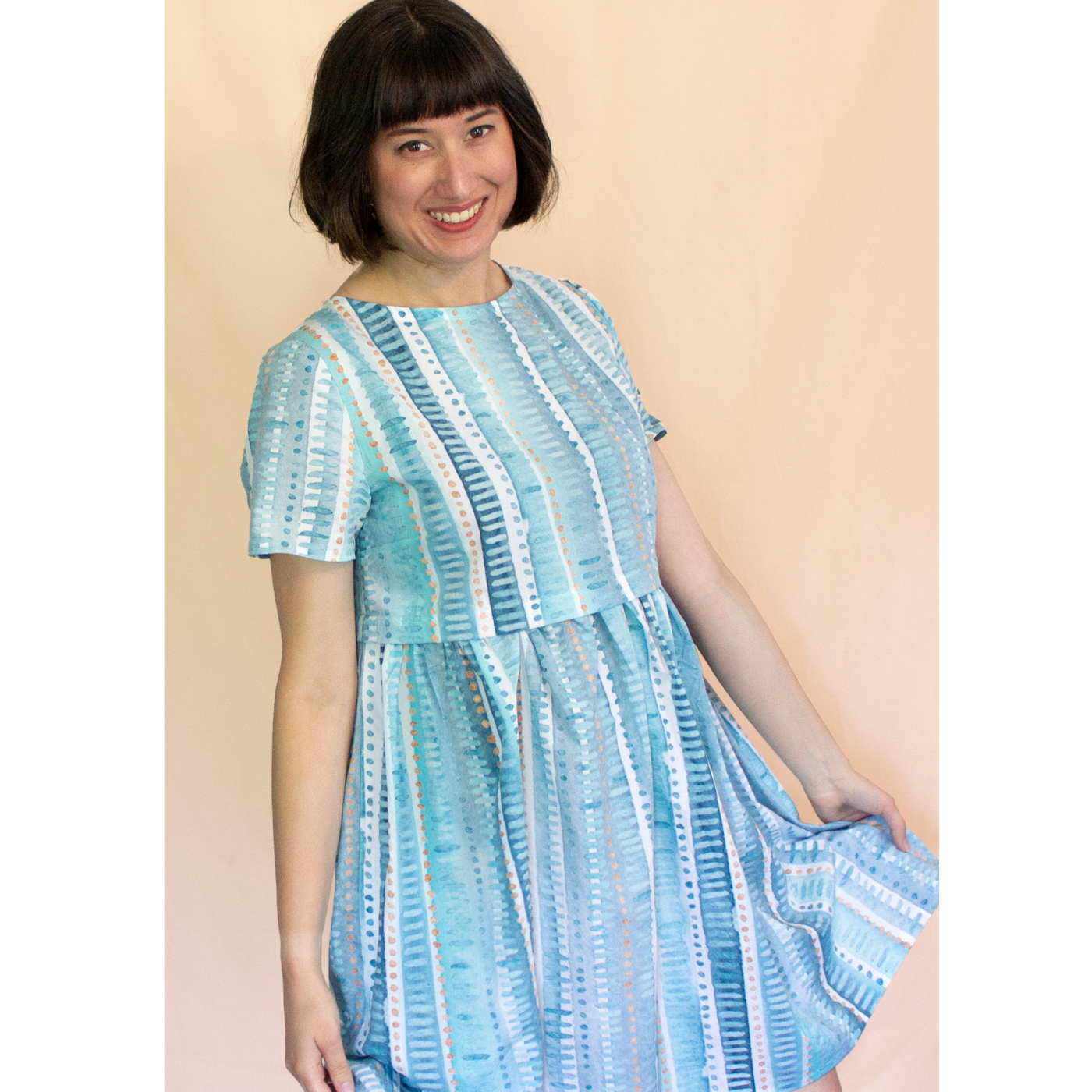 Emily smiles at the camera and stands in front of a peach wall wearing a knee-length dress with a print with long vertical strips in various shades of blue, gray and white and small horizontal lines running down the vertical strips in a differing blue, gray or white.