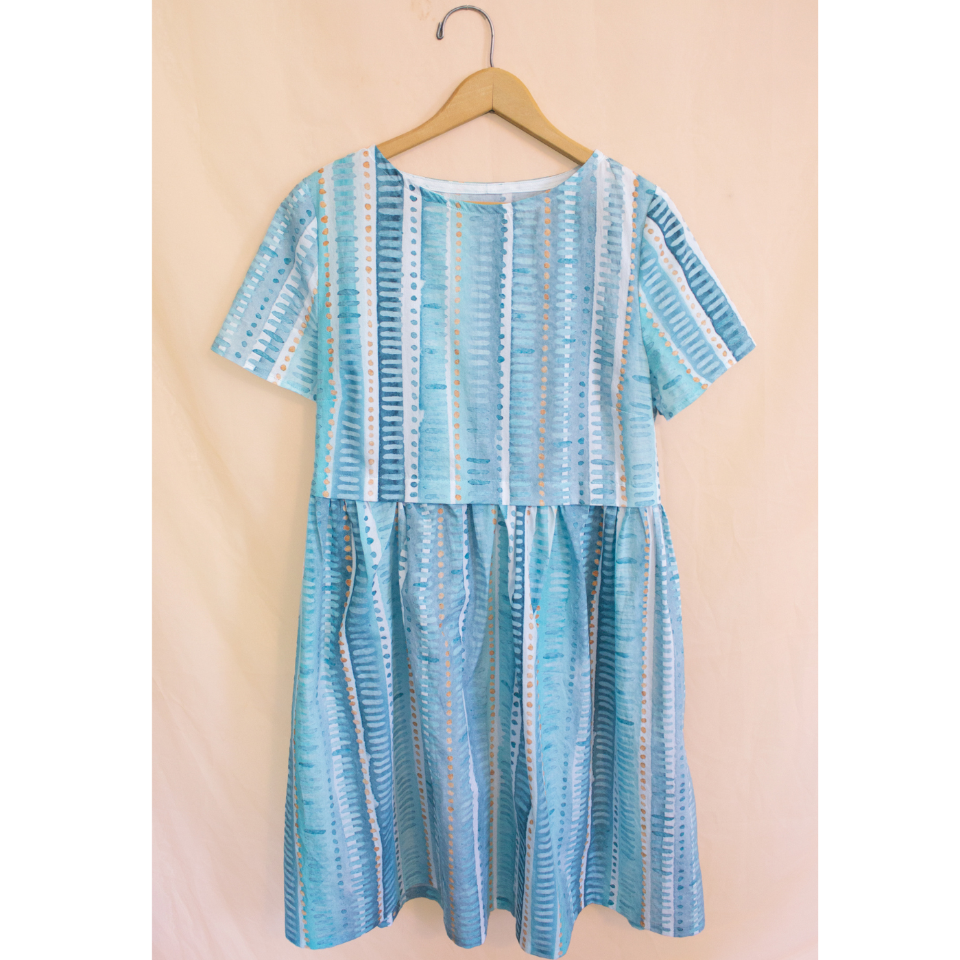 dress with a print with long vertical strips in various shades of blue, gray and white and small horizontal lines running down the vertical strips in a differing blue, gray or white shade hangs on a wooden hanger in front of a peach wall.