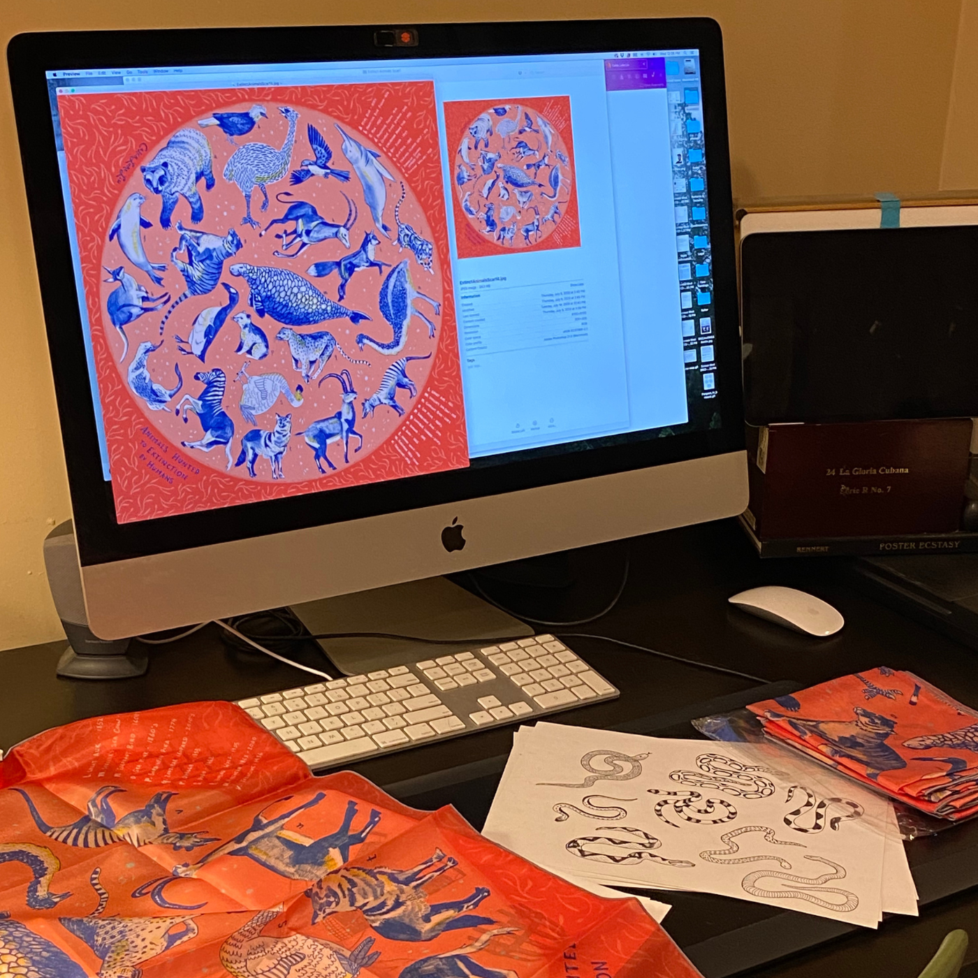 A design by Caleb Luke Lin is up on a computer monitor and also on a displayed scarf to the bottom left of the monitor and a folded scarf to the bottom right. The design has a red background and in a lighter red circle in the center of the design, a range of extinct animals, from birds to beasts to aquatic creatures. A drawing of different snakes is on a piece of paper in black ink by the computer’s keyboard.