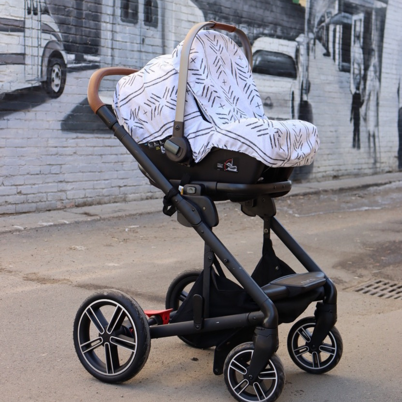 A black stroller sits in front of a black-and-white mural painted on a brick wall. The mural is of cars parked along a street with several pedestrians walking on a sidewalk to the right. A fabric cover is over the top of the stroller featuring a design with a white background and small black lines in geometric shapes.