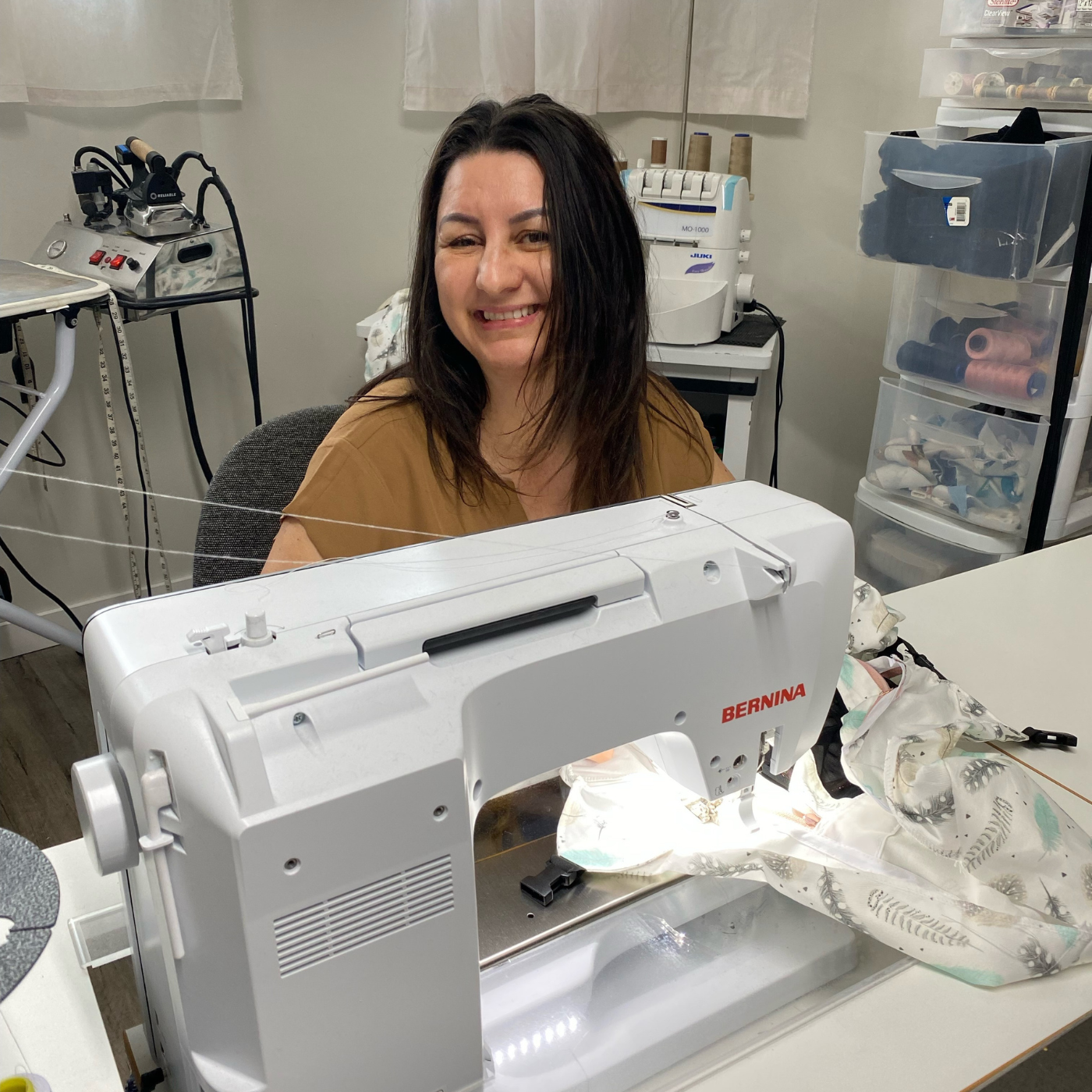 Elizabeth Waldner smiles at the camera from behind a sewing machine. White fabric with gray and mint leaves is on the table in front of the sewing machine.