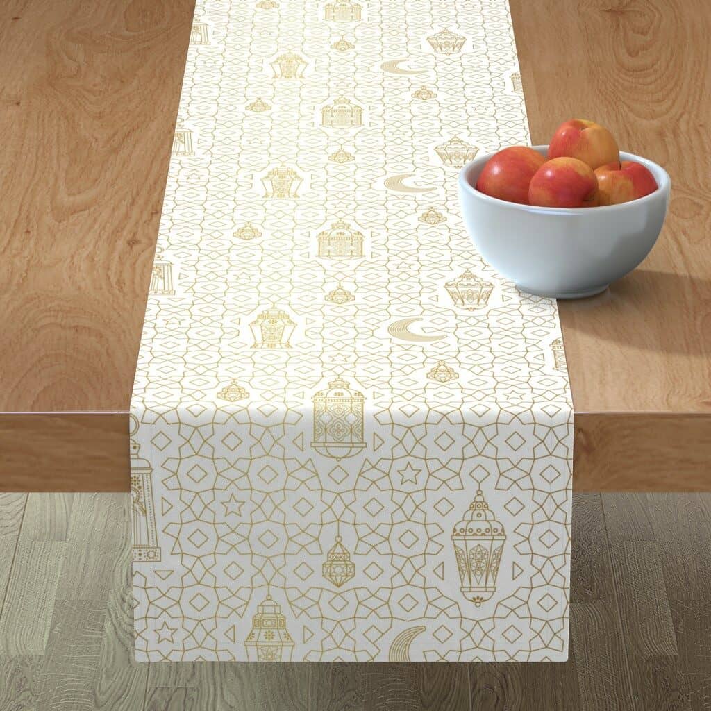 A table runner with a white background and gold Arabic lanterns of varying sizes amidst rows of stars lays on a wooden table. A white ceramic bowl full of red apples is to the photo's bottom right. 