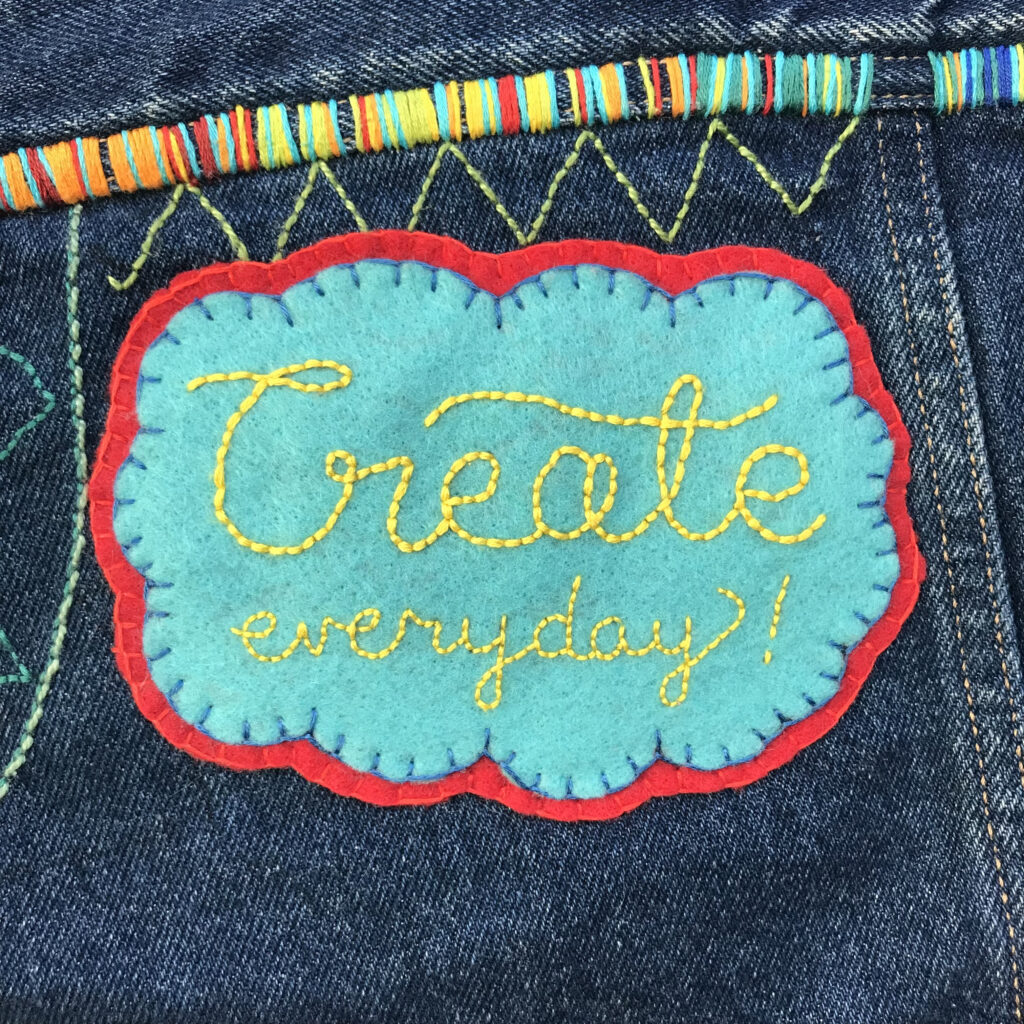  turquoise felt patch with a red felt rim and scalloped edges has the words “Create everyday!” embroidered in yellow. The patch is placed on denim with yellow zig zag stitches directly above it and a strip of small vertical stitches in a rainbow of colors. 