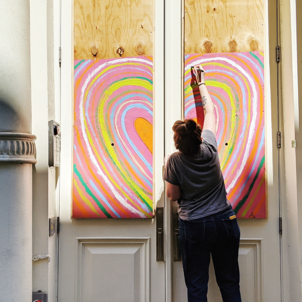 With her back to the camera, Jessie paints a large heart in orange, pink, yellow, green, white and blue on wooden boards placed on top of white shop doors.  