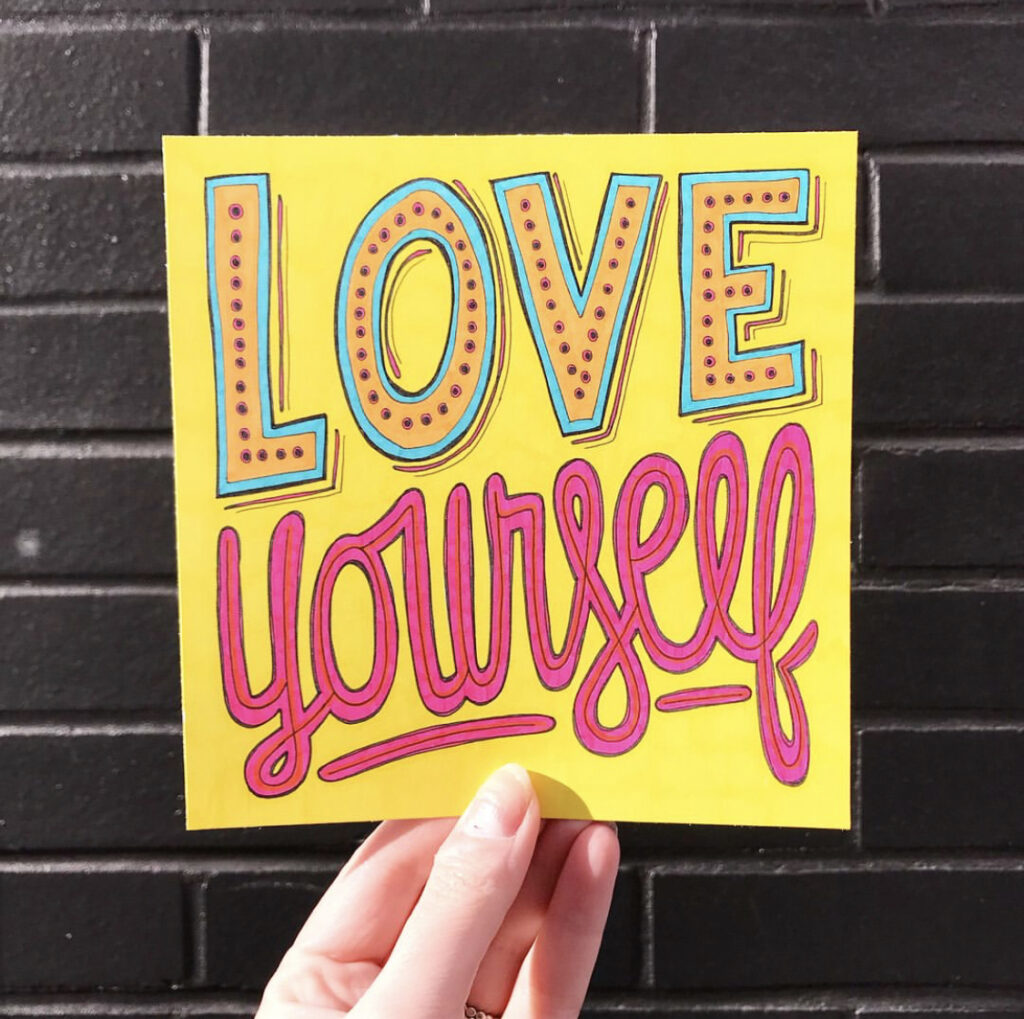 A hand holds up a square piece of yellow paper with the words “Love yourself” hand lettered. The word “love” is in all caps outlined in black and with an orange and turquoise center with black dots. The word “yourself” is in lowercase below it in magenta. 