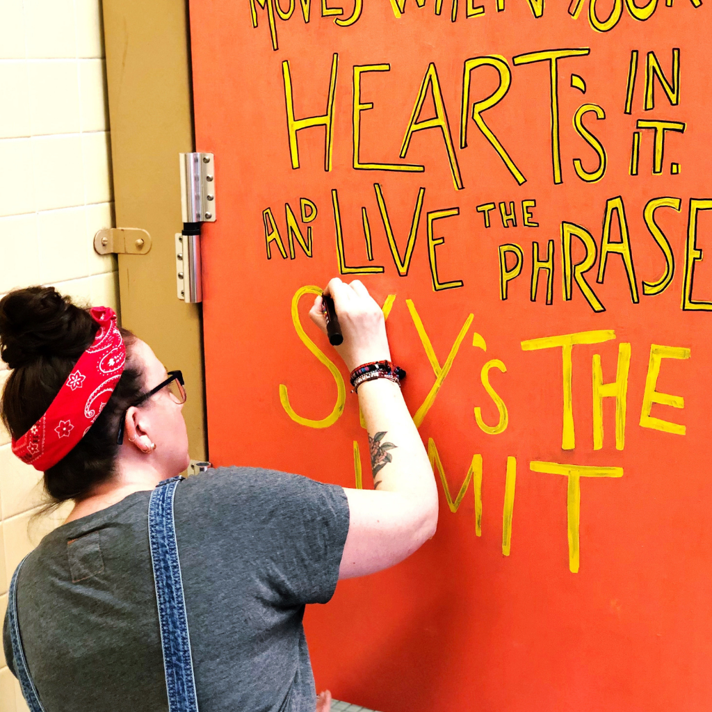 Jessie is working on a hand-letter mural in yellow, outlined in black ink on a bright orange bathroom stall door. The quote she is working on, “Only make moves when your heart's in it and live the phrase, "Sky's The Limit" is by The Notorious BIG and from the song Sky’s the Limit. 