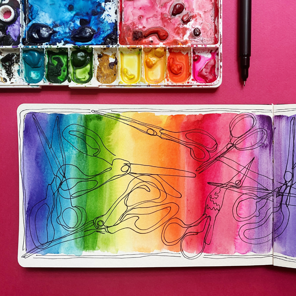 A watercolor palette is placed on a red surface with a black pen to its right. Below the palette is an open sketchbook painted with vertical stripes of rainbow colors from purple to blue, green, yellow, orange, red, pink and back to purple again. Scissors are drawn on top of the painted rainbow sections in black ink, pointing in all directions. 