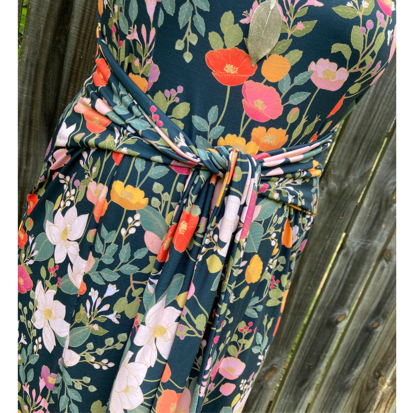 A close up of the mid-section of a dress with two thin fabric strips that tie in the front. The fabric has a black background and a floral print with orange, yellow and pink poppies and green flowers around them.