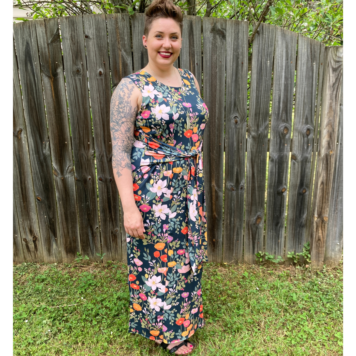 Jessica stands smiling at the camera wearing a dress that has two thin fabric strips that tie in the front. The fabric has a black background and a floral print with orange, yellow and pink poppies and green flowers around them. She is standing in a patch of grass near a tall wooden fence.