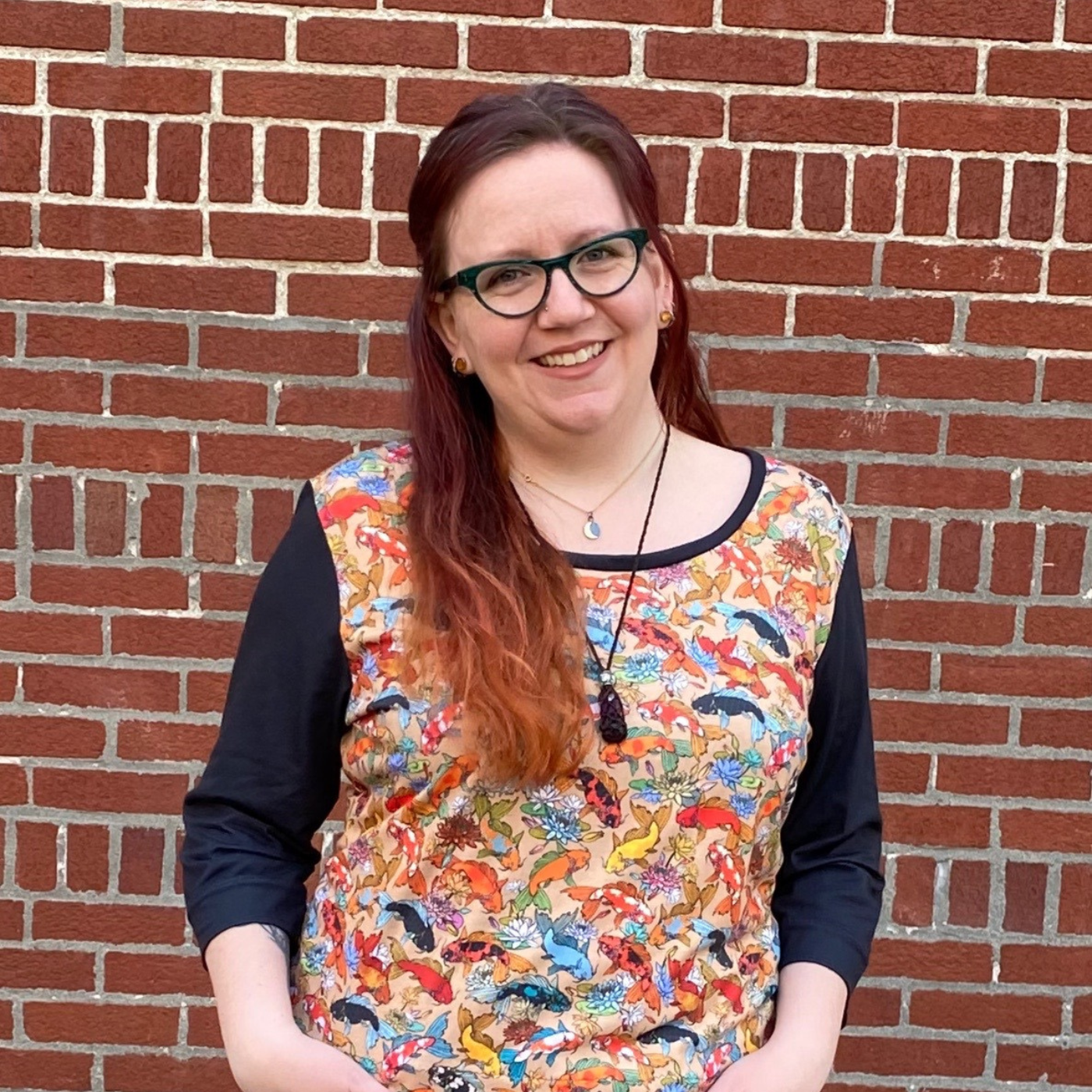 essie stands in front of a red brick building smiling, wearing a three-quarter-length-sleeve top with black sleeves. A print of rainbow koi swimming all over a peach background is all over the rest of the t-shirt except for a small bit of black fabric around the neck.