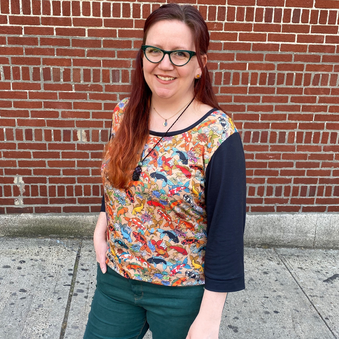 Jessie stands in front of a red brick building smiling, wearing a three-quarter-length-sleeve top with black sleeves and dark teal pants. A print of rainbow koi swimming all over a peach background is all over the rest of the t-shirt except for a small bit of black fabric around the neck.