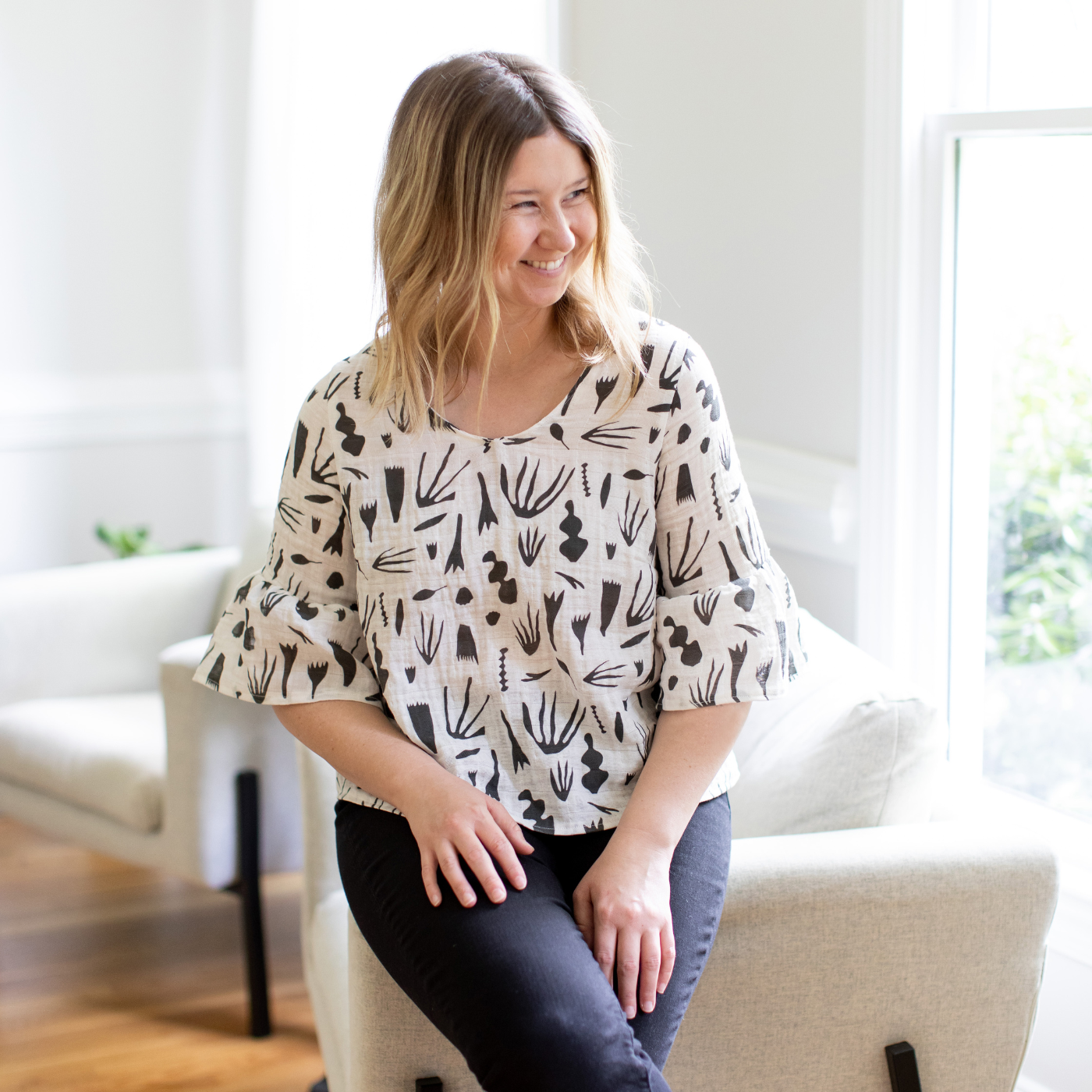 Katherine looks to the left and smiles, sitting on the arm of a white sofa. She wears a white top with ruffle sleeves and a black geometric pattern and black pants. Two large windows are to her left.