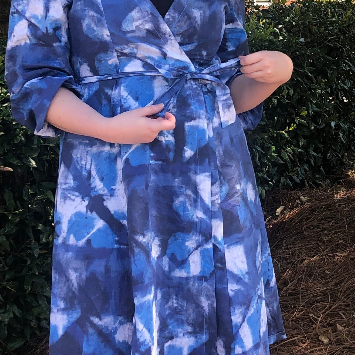 A close up of the midsection of the blue wrap dress Kelley is wearing with an indigo shibori design. Her hands are by the fabric bow tie at the waist which closes the dress.