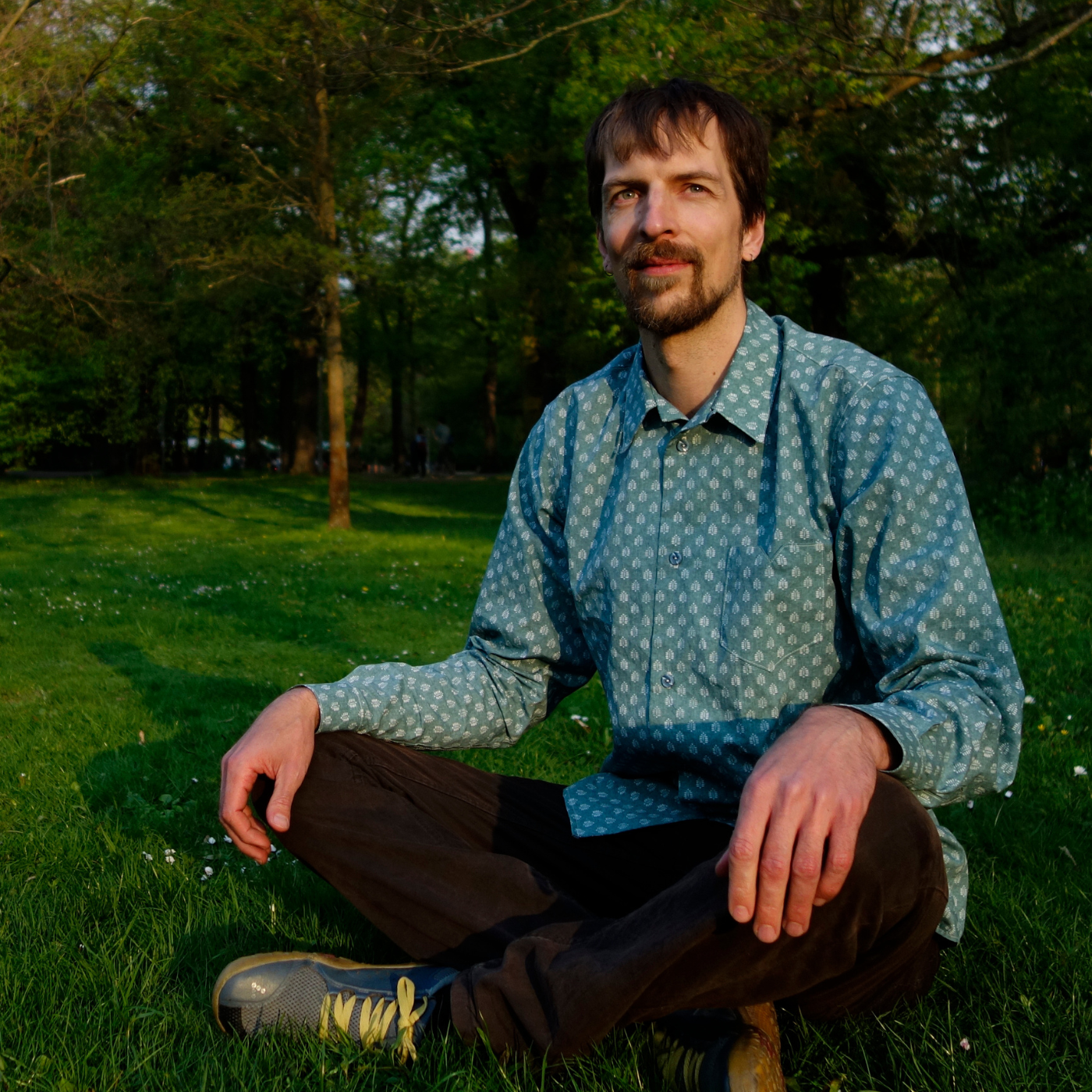 Kevin sits cross legged in a grassy park with his hands on his knees. He is wearing a teal button-up shirt and black pants and looking up and to the left. The shirt has repeating rows of small white block print ferns. There are tall trees behind him.