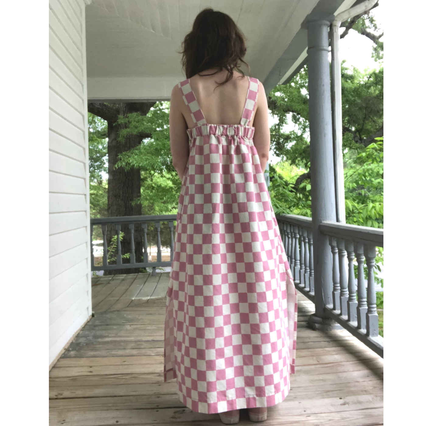 Lizard stands on a front porch with white walls, ceiling and railing and a wooden floor with their back to the camera. Their long maxi dress is down to their ankles and features a white-and-pink checkerboard design.