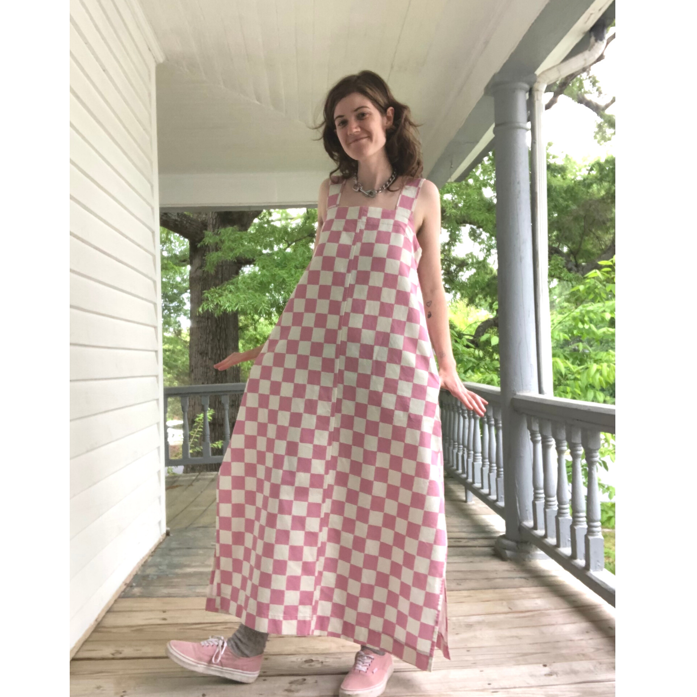 Lizard stands on a front porch with white walls, ceiling and railing and a wooden floor and is smiling at the camera. They are wearing a long maxi dress down to their ankles featuring a white-and-pink checkerboard design.