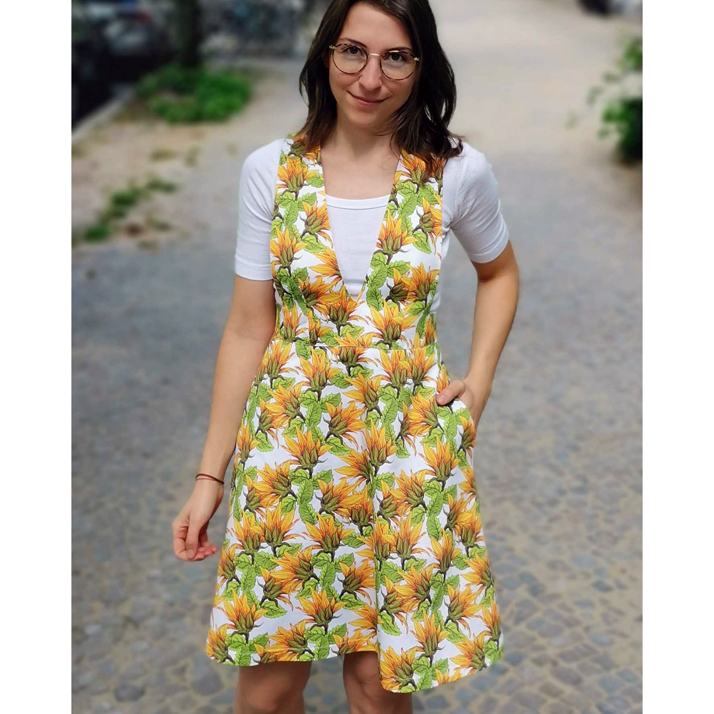 Maike stands looking at the camera with her left hand in her pocket. She is wearing a pinafore with a white background and sunflower design and a short-sleeve white shirt underneath.