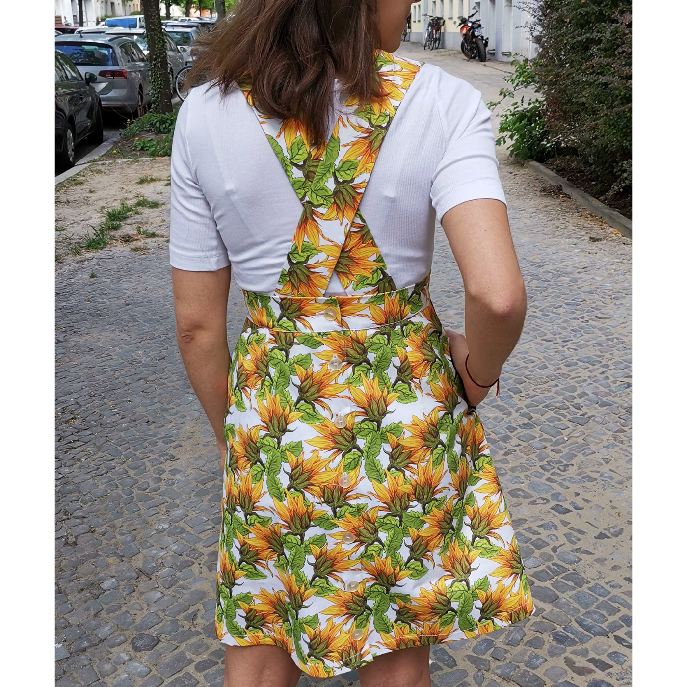 Maike stands with her back to the camera with her left hand in her pocket. She is wearing a pinafore with a white background and sunflower design and a short-sleeve white shirt underneath.