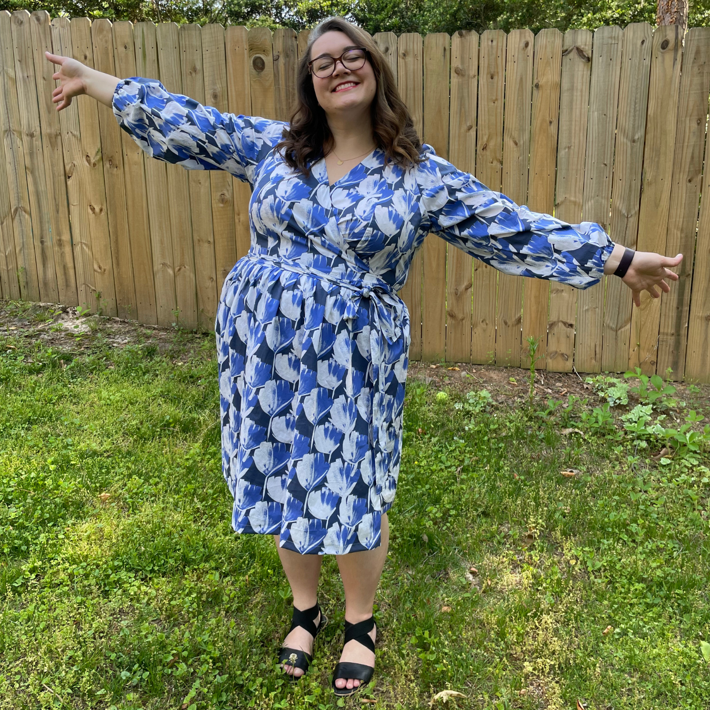 MaryAshlyn stands smiling at the camera holding both of her arms out straight. She is standing in the grass in front of a wooden fence and is wearing a dress with repeating white and blue flowers on a navy background.