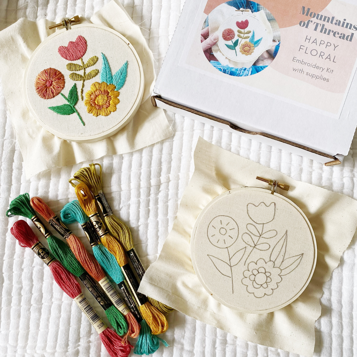 Items from Mountains of Thread’s “Happy Floral” embroidery kit are displayed on a white ribbed blanket. A box for the kit, showing a finished photo of a floral design and with “Mountains of Thread “Happy Floral” embroidery kit with supplies” typed on it in a dark gray font, is in the top right corner. A cream fabric square with a printed design of three large blooms with leaves is to the bottom right bound in a small round wooden embroidery hoop. Six full skeins of embroidery floss, from left to right, burgundy, sage, dusty orange, turquoise, goldenrod and yellowish green are grouped at the bottom left. A completed stitched floral design is at the top left of the photo.