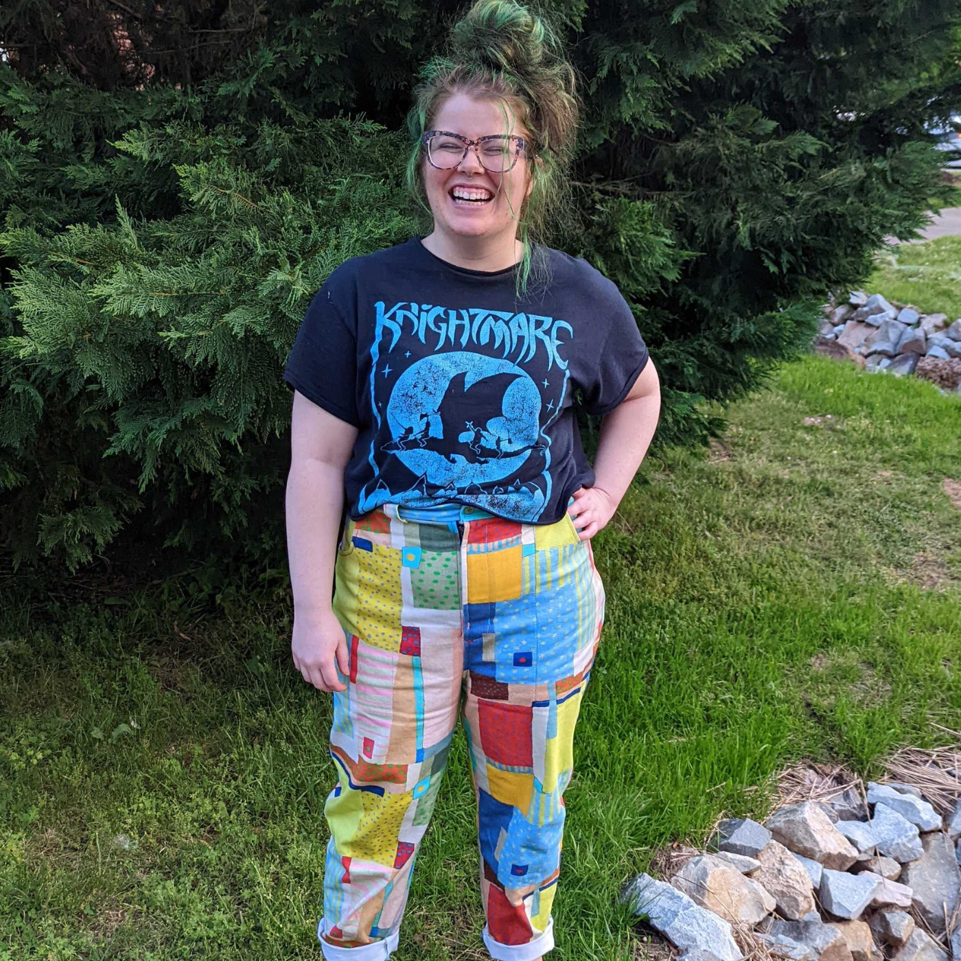 Nikki stands in front of the camera and is laughing. She is wearing a shirt for the band Knightmare and wearing pants featuring a design that looks like patchwork. Small squares and rectangles in varied sizes cover the design in yellow, red, blue, green and more Some blocks have stripes, others have dots. Both of her arms are held up in the air over her head. Her left leg is slightly in the air.