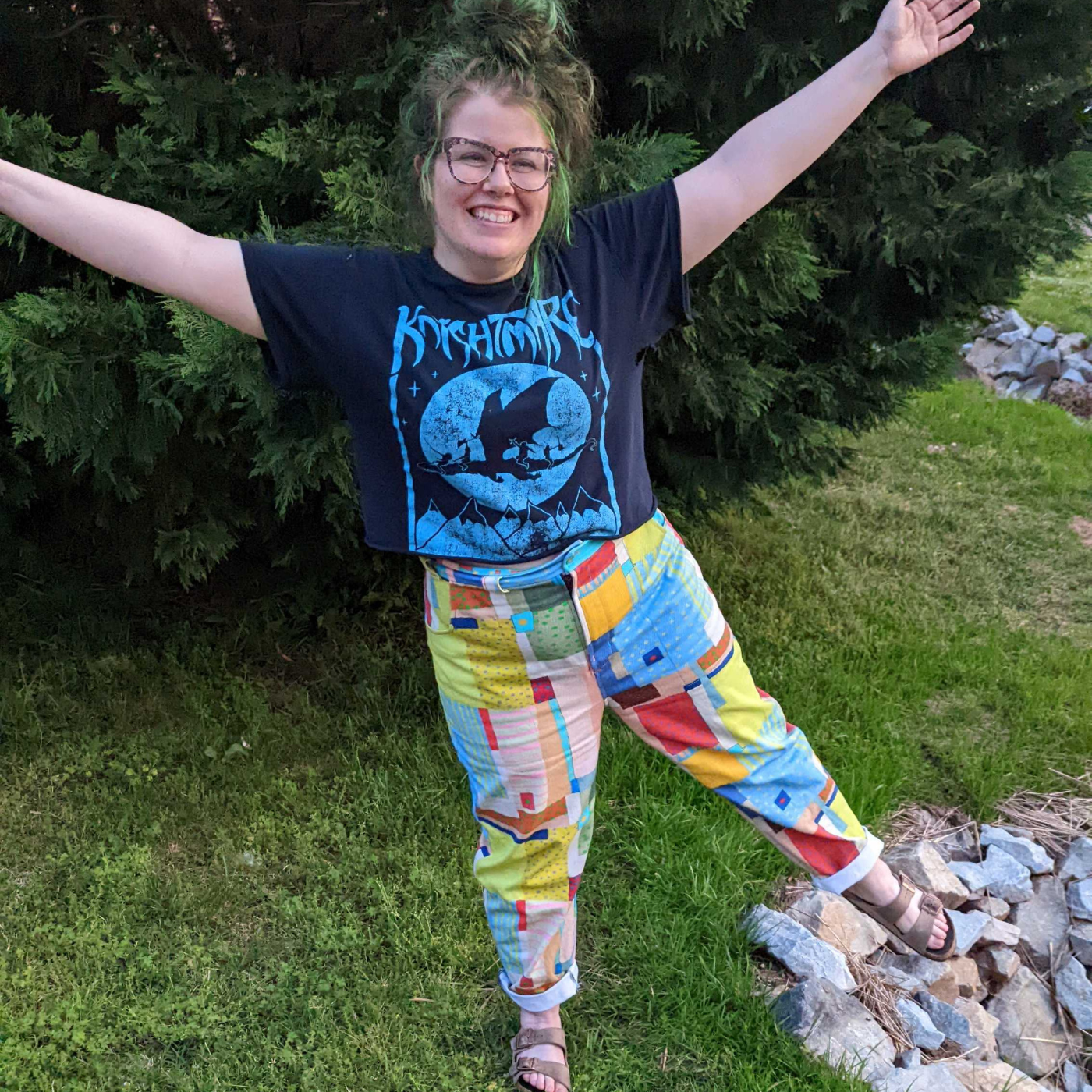 Nikki stands in front of the camera and is laughing. She is wearing a shirt for the band Knightmare and wearing pants featuring a design that looks like patchwork. Small squares and rectangles in varied sizes cover the design in yellow, red, blue, green and more Some blocks have stripes, others have dots. Her right hand is on her hip.