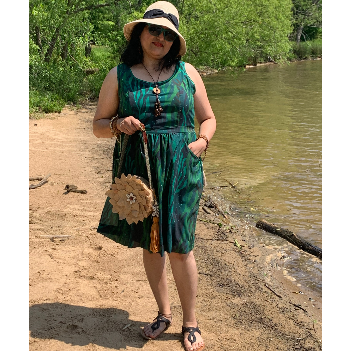 Nisha stands in the sand by a riverbank wearing a light brown sun hat and a sleeveless green dress featuring a design with black and green seaweed flowing in all directions. She is holding a brown floral purse and her left hand is in her pocket.