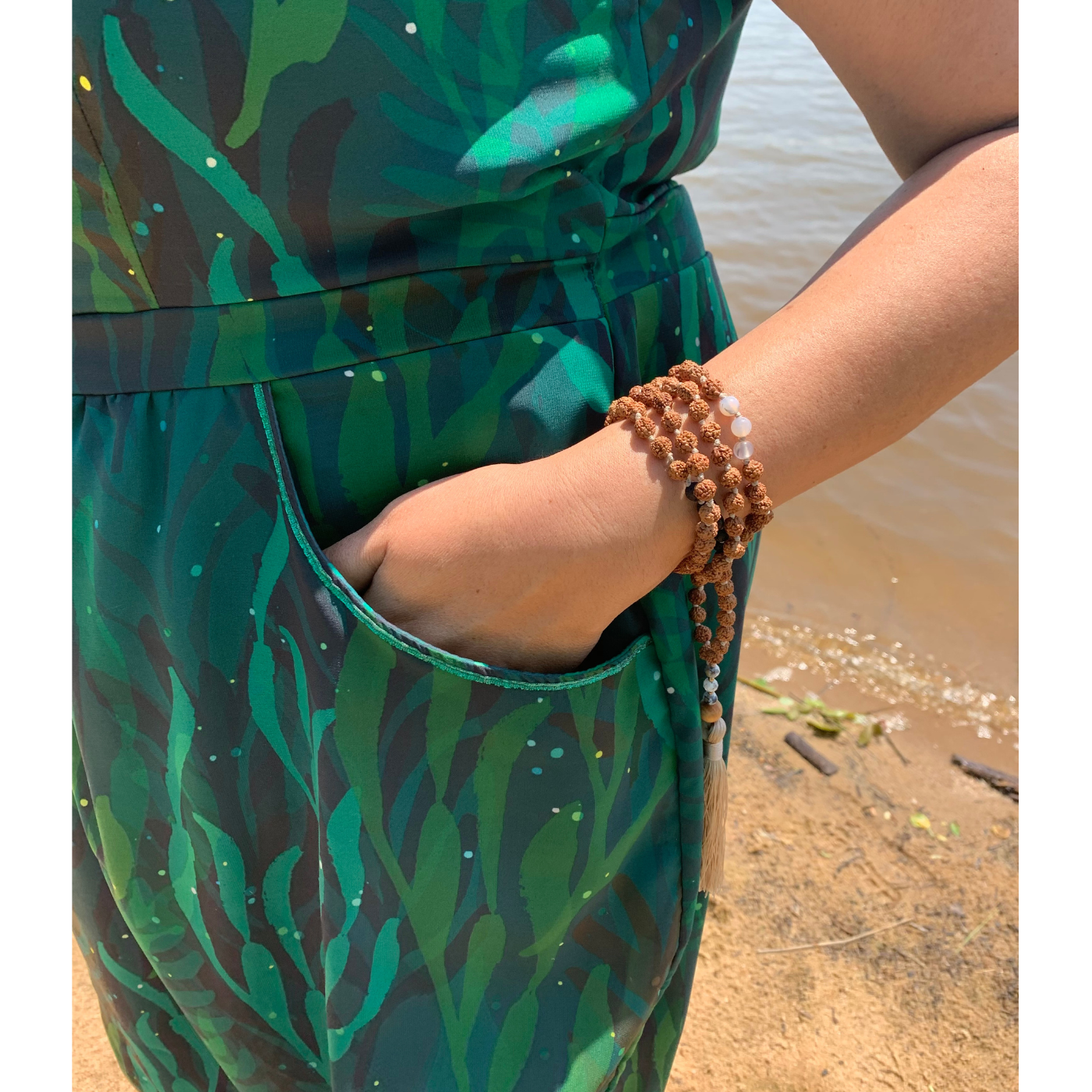 A close up of the midsection of a green dress Nisha is wearing featuring a design with black and green seaweed flowing in all directions. Her left hand is in her pocket and a bracelet with small brown round beads is on her wrist.
