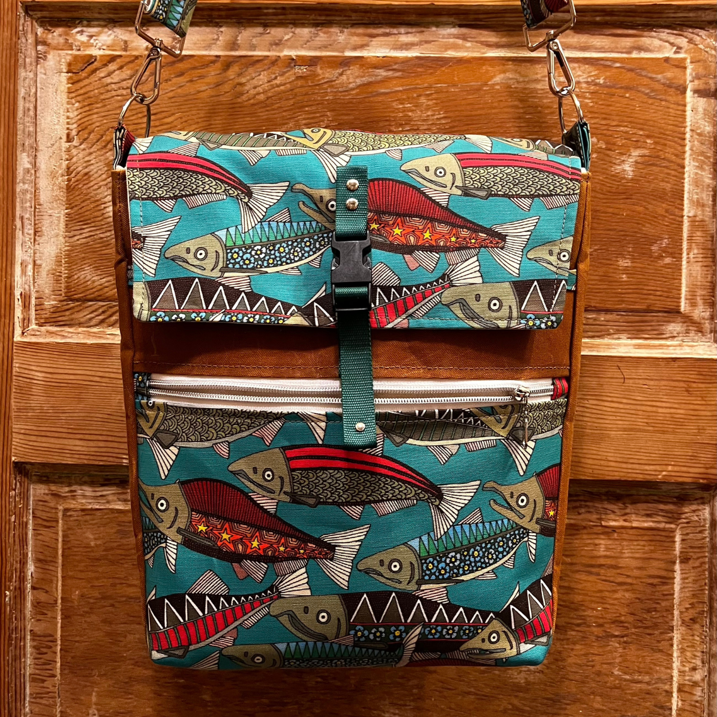 A handmade rectangular bag with a fish design (with a turquoise background and red, green, and blue fish, some going to the left, some going to the right) hangs against a brown wooden door. A buckle clasp closes the bag in the middle of the top third