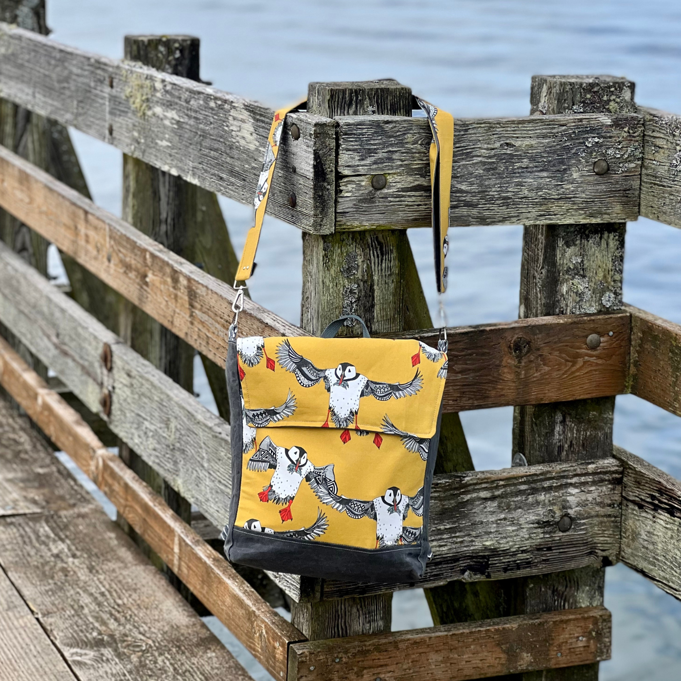 A handmade rectangular bag with a fish design (with a yellow background and rows of large white puffins with black-and-white wings outstretched and orange feet) hangs against a brown slatted fence on the edge of a pier overlooking the ocean. The bag has a top flap that closes in the top third.