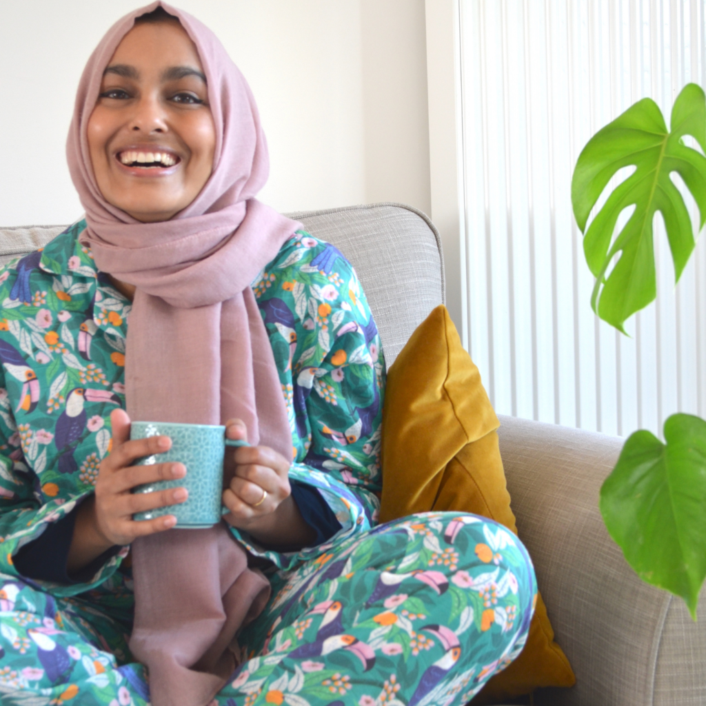 Rumana sits on a gray couch holding a mug in her hands, looking at the camera and smiling. She is wearing a dusty pink hijab and pajamas with a tropical print featuring toucans, pink and orange flowers and a bright blue background. To the right are a few monstera leaves. 