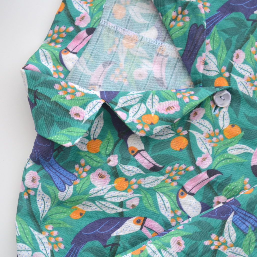 A close up of the top and neckline of pajamas with a tropical print featuring toucans, pink and orange flowers and a bright blue background.